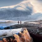 Two people on some rocks facing a huge wave