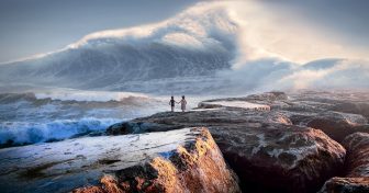 Two people on some rocks facing a huge wave