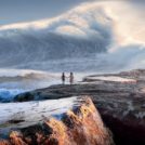 Two people on some rocks facing a huge wave